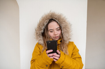 Happy young woman with Down syndrome weraing parka, leaning the wall and using smartphone. © Halfpoint