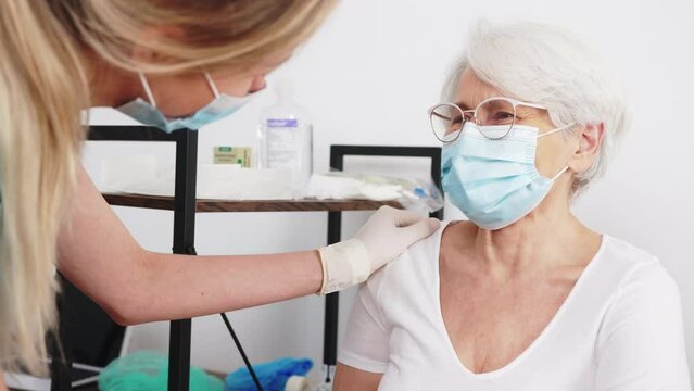 Indoor portrait of beautiful elderly woman in protective face mask talking to her nurse during health check-up. High quality 4k footage