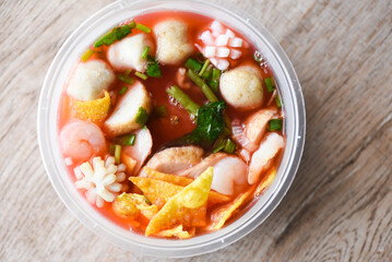 Red soup noodle pork balls crispy wontons with seafood squid shrimp and fish balls vegetable in soup bowl plastic on wooden table, top view Thai and China Asian food delivery concept