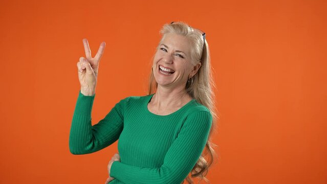Portrait of blonde happy mature woman giving peace victory sign 50s casual clothes look camera isolated on orange background with copy space studio.