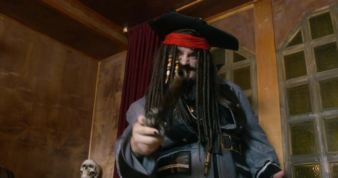 Crazy pirate captain in cocked hat with dark long braids points revolver standing against skull ON rack in ship cabin at voyage