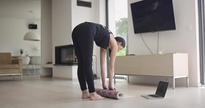 woman in sportswear doing stretching exercises while watching yoga training class on computer laptop online. Healthy girl exercising in living room with sofa couch in the background..