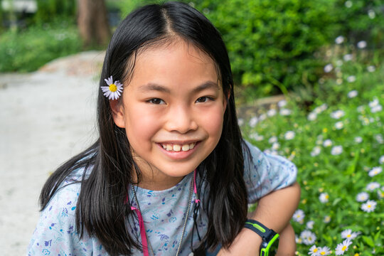 Portrait of Asian little girl with a cute smiling face, black long hair, enjoy in a garden, small amount of sweat on face, eyes looking at camera, small white flower at ear, close up image