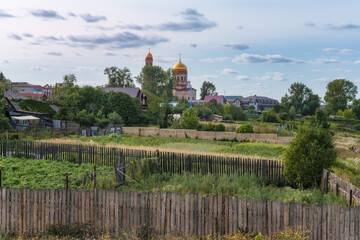 Fototapeta na wymiar Panorama of the village with the golden domes of the old church, wooden houses and gardens of the inhabitants, immersed in the greenery of trees and grass. Beautiful cloudy sky. Russia, Ural 