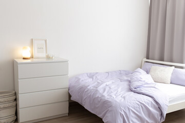 Bedroom in lilac color. Bed with purple linen, white chest of drawers, night salt lamp, frame, candles, grey curtains. Bedroom decor. Modern stylish interior.