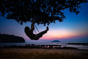 A silhouette woman plating swing on the beach, tropical sunset view of Thailand.