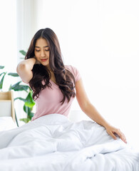 Obraz na płótnie Canvas Asian young beautiful happy sleepy female teenager in casual pajama outfit waking up fresh in morning sitting smiling closed eyes under white blanket on bed in bedroom massaging stretching neck