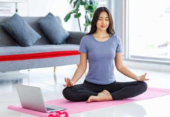 Asian young happy peaceful calm female model in casual sporty outfit sitting crossed legs in lotus position on yoga mat learning studying online meditation class via laptop computer in living room - obrazy, fototapety, plakaty