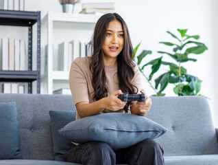 Asian young beautiful cheerful happy exciting female gamer model sitting on cozy couch smiling holding joystick controller playing fun game via console gaming platform online in living room at home