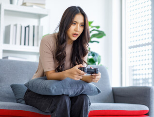 Obraz premium Asian young beautiful cheerful happy exciting female gamer model sitting on cozy couch smiling holding joystick controller playing fun game via console gaming platform online in living room at home