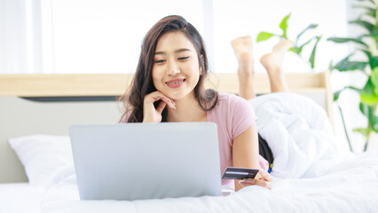 Obraz na płótnie Canvas Asian young happy cheerful beautiful female model in casual pajama outfit lying down smiling hold credit card on bed in bedroom browsing surfing internet shopping online via laptop notebook computer