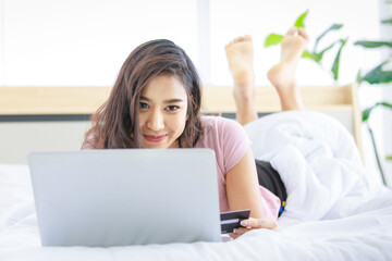 Asian young happy cheerful beautiful female model in casual pajama outfit lying down smiling hold credit card on bed in bedroom browsing surfing internet shopping online via laptop notebook computer