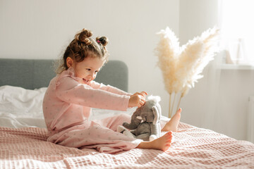 A cheerful little girl is sitting on the bed after waking up and playing with her toy elephant and laughing