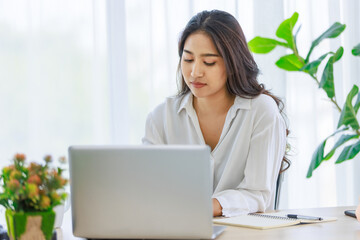 Asian young beautiful professional successful smart female businesswoman working with data information on laptop computer writing note on notebook at workstation desk.