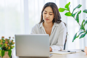 Asian young beautiful professional successful smart female businesswoman working with data information on laptop computer writing note on notebook at workstation desk.