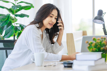 Obraz premium Asian young beautiful professional successful smart female businesswoman on call via smartphone while working with data information on laptop computer writing note on notebook at workstation desk