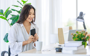 Asian young beautiful professional successful smart female businesswoman on call via smartphone while working with data information on laptop computer writing note on notebook at workstation desk