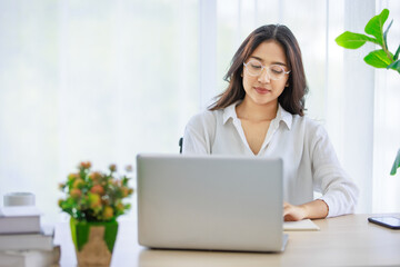 Asian young beautiful female university student wearing optical eyeglasses sitting at working desk remotely studying learning streaming online via laptop notebook computer at home during quarantine