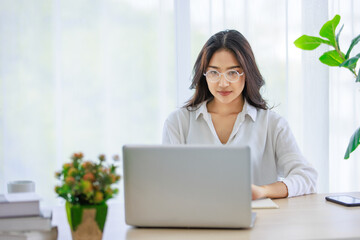 Asian young beautiful female university student wearing optical eyeglasses sitting at working desk remotely studying learning streaming online via laptop notebook computer at home during quarantine