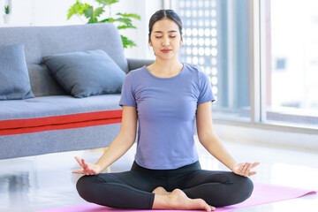 Asian young happy peaceful calm female model in casual sporty outfit sitting crossed legs in lotus position on yoga mat learning studying online meditation class via laptop computer in living room - obrazy, fototapety, plakaty