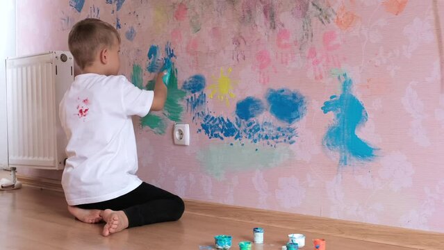 A little boy draws with his palms and paints on the pink wallpaper on the wall in his room. Happy Childhood Concept.