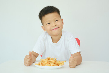 Cute happy smiling Asian 5 yers old kindergarten boy child eating French fries potato chips sitting at the table Isolated over white wall background, Unhealthy Kids Foods concept