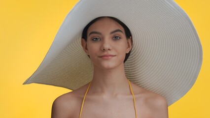 Brown-haired beauty model in a big white hat looks at camera against yellow background | Sun protection and body care commercial