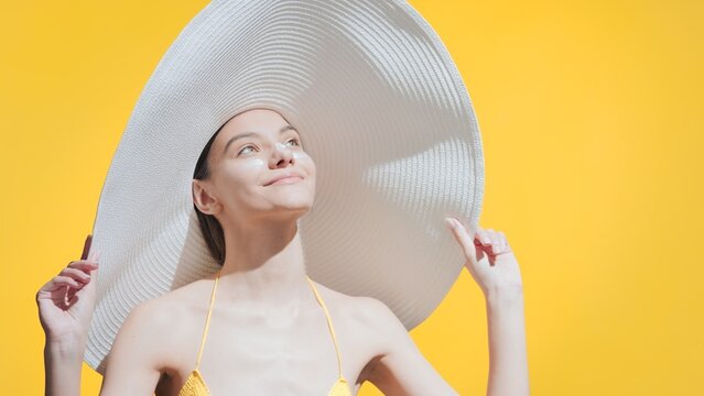 Young Gorgeous Brunette Caucasian Woman In A Big White Hat With Sunblock On Her Cheeks And Nose Enjoys The Sun Smiling Against Yellow Background | Sunscreen Commercial Concept