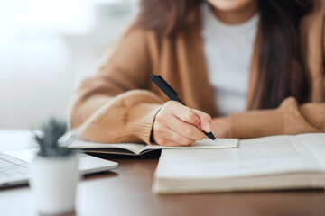 Close up - Hand of woman writing in spiral notepad placed on wooden desktop with various items at...