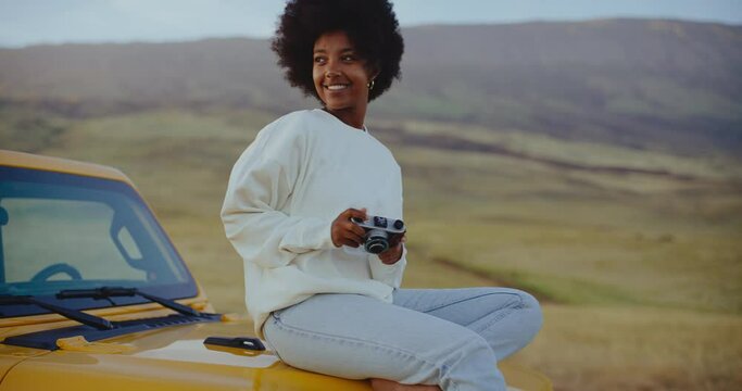 Beautiful young black african american woman relaxing and taking pictures at sunset with vintage camera on epic road trip, freedom and travel concept