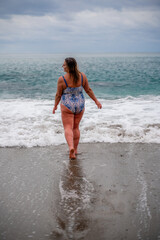A plump woman in a bathing suit enters the water during the surf. Alone on the beach, Gray sky in the clouds, swimming in winter.