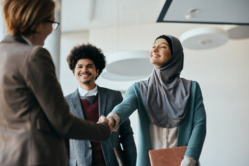 Happy Muslim businesswoman shaking hands with female colleague in hallway of an office building.