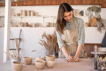 Young beautiful woman with long hair in an apron creates a handmade ceramic bowl from clay. Creative workshop. The concept of skill and entrepreneurship.