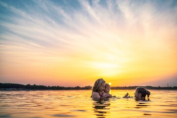 Mom with the baby she holds in her arms playing with her daughter and splashing in the lake against background of sunset