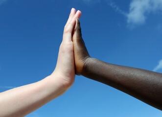 a white-skinned hand and a black-skinned hand high-five each other in front of a blue sky