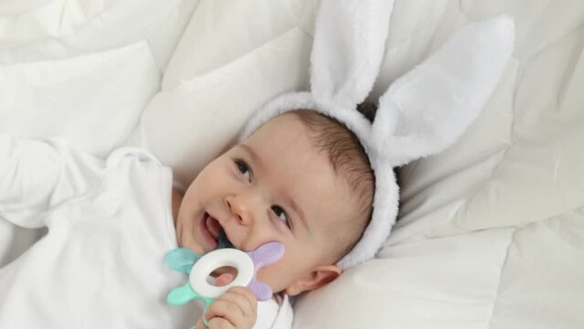 cute little boy , baby, toddler, sitting on white blanket with white bunny easter ears and chewing a soft teething toy. adorable photography for easter holidays. rubber toy for child’s teeth.