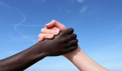 a white-skinned hand and a black-skinned hand hold each other in front of a blue sky
