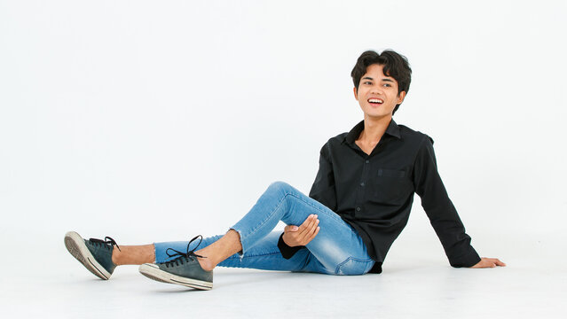 Portrait isolated cutout studio shot of Asian young LGBTQ gay glamour male model in casual black shirt and jeans outfit sitting stretching posing gesturing on floor look at camera on white background