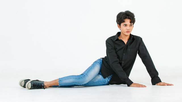 Portrait isolated cutout studio shot of Asian young LGBTQ gay glamour male model in casual black shirt and jeans outfit sitting stretching posing gesturing on floor look at camera on white background
