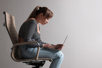 Young woman with poor posture using laptop while sitting on chair against grey background, space...