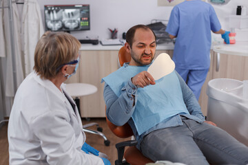 Fototapeta premium Patient holding mirror looking at teeth procedure after dental surgery during stomatology examination in stomatological office room. Dentist senior woman explaining oral hygiene. Concept of medicine
