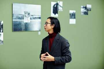 Serious professional art critic wearing eyeglasses standing in modern art gallery looking at black and white photos on wall at exhibition