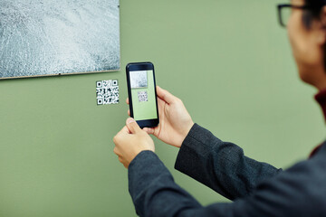 Unrecognizable young man spending time in modern art gallery scanning QR code using his smartphone to get more information about photo on wall - Powered by Adobe