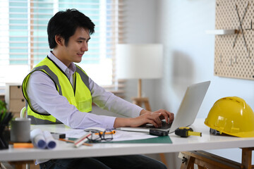 Handsome male construction engineer in white shirt and yellow vest using laptop computer and working with blueprints.