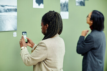 Young African American woman attending exhibition in modern art gallery scanning QR code using...