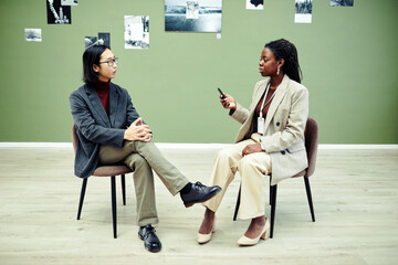 African American woman sitting on chair holding voice recorder interviewing modern young Asian...