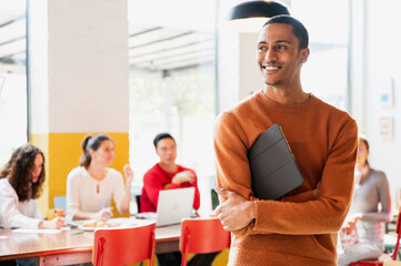 Attractive Moroccan young male in casual clothes standing and looking at his side smiling while holding a tablet. Teamwork sitting at desk on background.