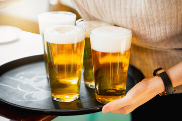 Unrecognizable waitress serving three glasses with iced beer with foam on a tray.