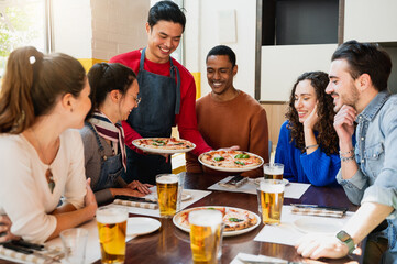 Multiracial group of friends sitting at table restaurant looking hungry the pizzas that the waiter is bringing.