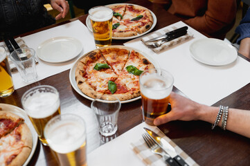 Top view setting table restaurant with Italian traditional pizza margherita and glasses of beer. Unrecognizable hands grabbing mug.